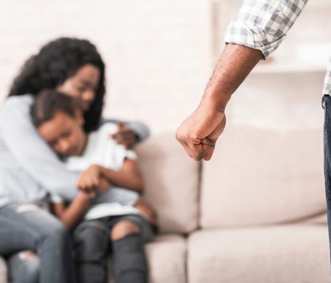 Mother protecting child on couch while facing an aggressive partner during a domestic violence incident.